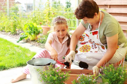A woman and a young girl are engaged in gardening within a well-maintained outdoor space that appears to be part of a residential backyard near Thornton Heath. The woman, dressed in a light green top and wearing a floral-patterned apron, is helping the girl plant or tend to flowers in a long, rectangular flower bed made of wood. The girl, wearing a pink shirt with white and pink striped trousers, is focused on her task, holding a small gardening tool and working with soil in the plant bed. In the foreground, there are vibrant green plants and blooming flowers, while in the background, a lush garden area with a variety of shrubs, small trees, and potted plants is visible. The garden features a neatly trimmed lawn and a paved patio area that complements the overall landscaped environment. The weather appears sunny and clear, casting natural light on the scene, which reflects regular outdoor maintenance and gardening care typical of services offered by Gardening Services Thornton Heath. The scene captures a moment of outdoor family gardening, highlighting a tidy and inviting garden space with healthy plants, well-organized flower beds, and a peaceful green environment.