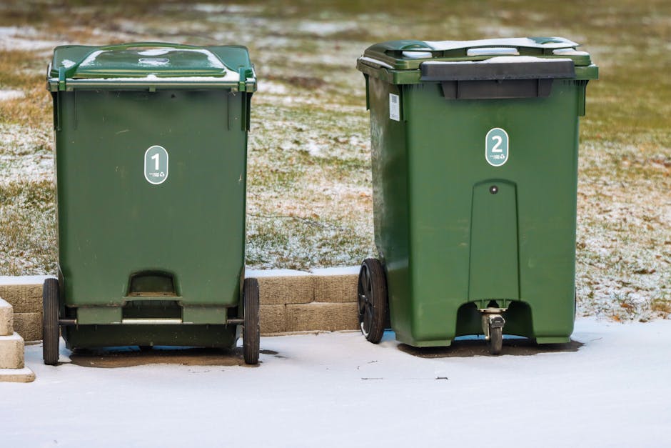 Two green outdoor wheelie bins placed side by side on a snowy pavement in a garden setting; the left bin is numbered '1' and appears slightly more weathered, while the right bin, numbered '2', has a l