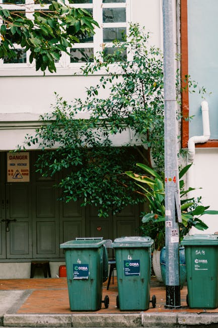 Two green outdoor wheelie bins placed side by side on a snowy pavement in a garden setting; the left bin is numbered '1' and appears slightly more weathered, while the right bin, numbered '2', has a l