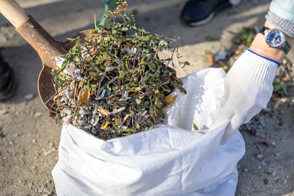 A person wearing a white gardening glove and a blue wristwatch is using a small shovel to scoop up a pile of garden debris, including branches, leaves, and small twigs, into a white garden waste bag p